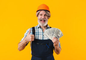 Picture of emotional positive happy elderly grey-haired bearded man builder in helmet posing isolated over yellow wall background holding money.